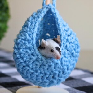 May include: A small, black and white mouse resting inside a blue crocheted hanging bed.