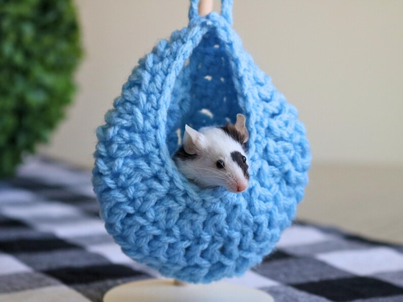 May include: A small, black and white mouse resting inside a blue crocheted hanging bed.
