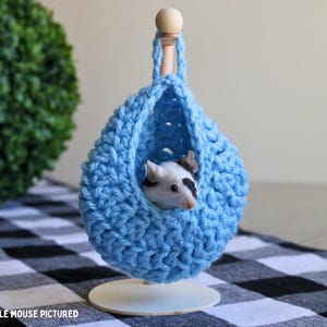 May include: A blue crocheted hanging bed for small pets, such as mice or hamsters. The bed is suspended from a wooden dowel and has a small mouse inside.