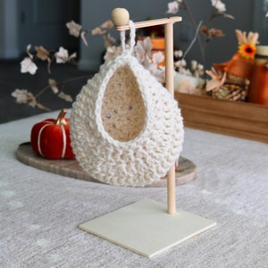 May include: A white crocheted hanging basket with a wooden stand. The basket is shaped like a teardrop and has a wide opening at the top. The stand is made of natural wood and has a square base.