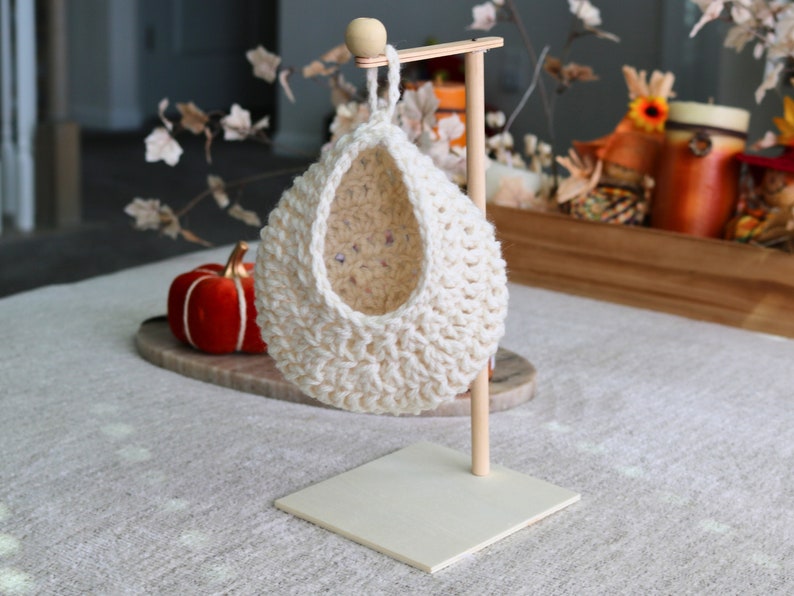 May include: A white crocheted hanging basket with a wooden stand. The basket is shaped like a teardrop and has a wide opening at the top. The stand is made of natural wood and has a square base.