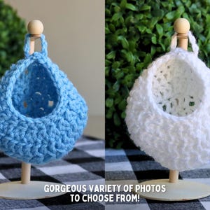 May include: Two crocheted hanging baskets, one blue and one white, are displayed on wooden stands. The baskets are made with a textured stitch and have a round shape.