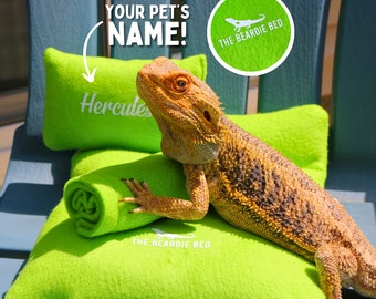 May include: A green, fleece-lined bed for bearded dragons, with the words "The Beardie Bed" embroidered on it. A bearded dragon named Hercules is lying on the bed.