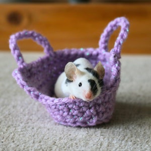 May include: A small black and white mouse sits in a purple crocheted basket with handles. The basket is on a light brown carpet.