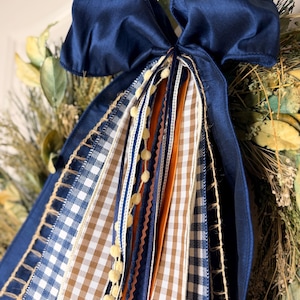 May include: A decorative bow featuring a large navy blue satin ribbon at the top with multiple hanging streamers. The streamers include navy and white gingham, tan and white gingham, solid orange, and cream pom-pom trim ribbons.