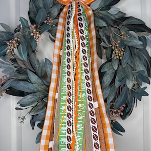 May include: A decorative wreath with a bow made of orange and white checkered ribbon. The wreath features a variety of ribbons with football and yard line designs. The wreath is adorned with green leaves and small berries.