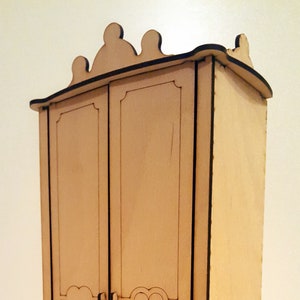 May include: A wooden dollhouse wardrobe with a decorative top and two doors. The wardrobe is made of light brown wood and has a simple design.