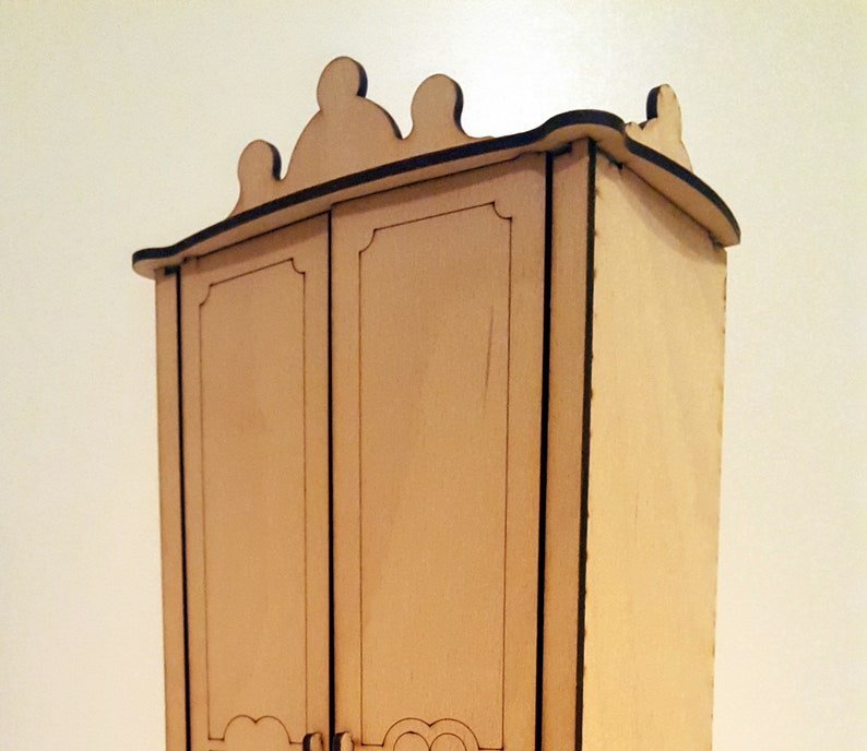 May include: A wooden dollhouse wardrobe with a decorative top and two doors. The wardrobe is made of light brown wood and has a simple design.