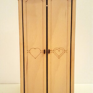May include: A wooden dollhouse wardrobe with two doors, each featuring a heart-shaped cutout. The wardrobe is painted in a light wood color and has a decorative top.