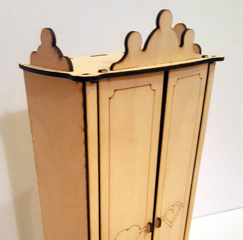 May include: A wooden dollhouse wardrobe with two doors and a decorative crown-shaped top. The wardrobe is made of light brown wood and has black trim.