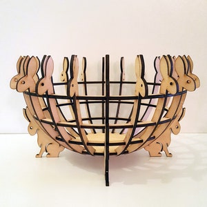 Op de afbeelding: Een houten kom in de vorm van een mand met konijnen silhouetten rond de rand. De kom is gemaakt van lichtbruin hout met donkerbruine accenten.