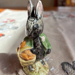 May include: A ceramic figurine of a rabbit, wearing a green jacket and holding a basket. The rabbit has black and gray fur with pink-lined ears. The basket contains orange-colored items. The figurine is on a beige surface with embroidered details.