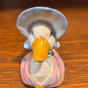 May include: A ceramic duck figurine wearing a blue bonnet and a pink and blue dress. The duck has a bright orange beak and is set against a wooden background. The figurine is a decorative item.