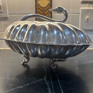 May include: A silver-colored serving dish with a swan-shaped handle. The dish has a ribbed, oval design and rests on three curved feet. The background features a tiled wall and a dark countertop.