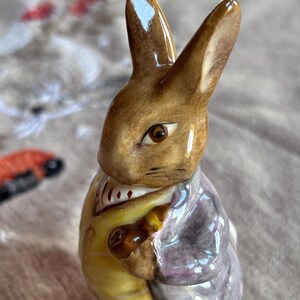 May include: A ceramic figurine of a rabbit, featuring a brown and tan head with long ears. The rabbit wears a yellow coat, a purple jacket, and a white shirt with a red and white pattern. The figurine is a collectible item.