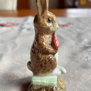 May include: A ceramic figurine of a brown rabbit holding a red carrot. The rabbit is sitting on a green bench atop a bed of straw. The figurine has detailed fur and is a collectible item.