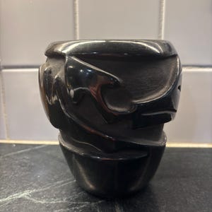 May include: A black, carved vessel with a textured surface. The vessel has a rounded base and a wider top, with carved, abstract designs around the sides. The object is made of a dark, glossy material.