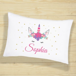 Puede incluir: Almohada blanca con un diseño de unicornio con una corona de flores rosas y un cuerno morado. El unicornio tiene los ojos cerrados y el nombre "Sophia" está escrito en rosa debajo del unicornio.
