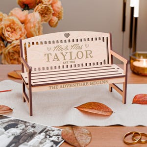 Könnte beinhalten: Eine Holzbank mit den Worten "Mr & Mrs TAYLOR EST. 2026" und "THE ADVENTURE BEGINS" eingraviert. Die Bank hat einen herzförmigen Ausschnitt und ist braun und beige. Es ist ein Hochzeits- oder Jubiläumsgeschenk.