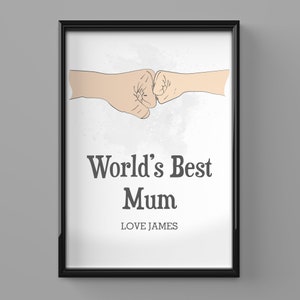 Puede incluir: Impresión enmarcada en negro con el texto "World's Best Mum" y "Love James". La imagen muestra dos puños chocando, con un fondo de estilo acuarela gris claro. El texto y la imagen son de color gris oscuro.