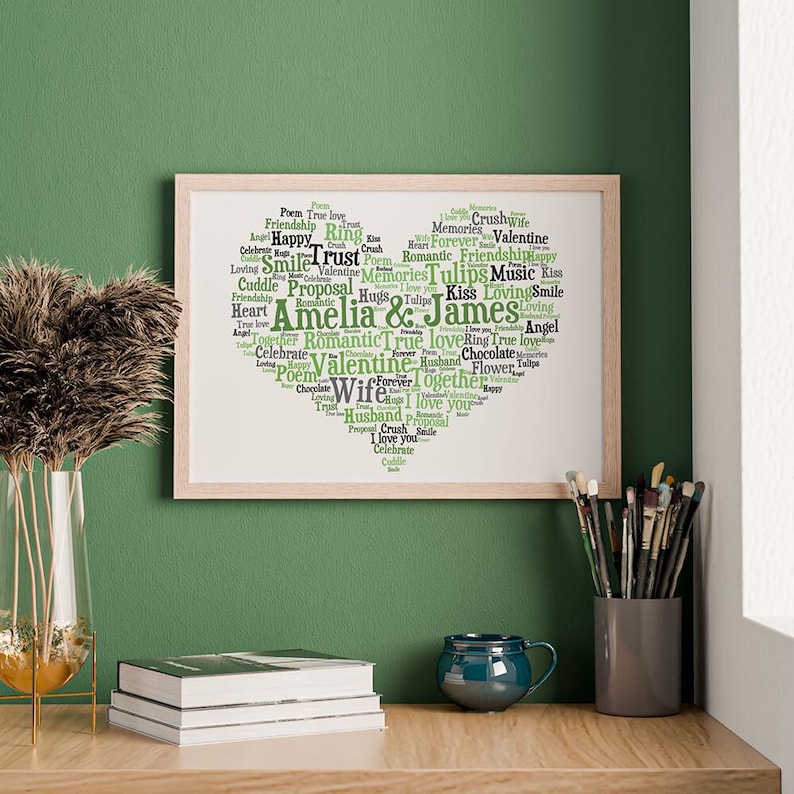 Puede incluir: Una impresi&oacute;n rectangular enmarcada que presenta una nube de palabras en forma de coraz&oacute;n compuesta por varios t&eacute;rminos rom&aacute;nticos y los nombres Amelia & James en tonos verdes y negros sobre un fondo blanco, en un marco de madera clara.