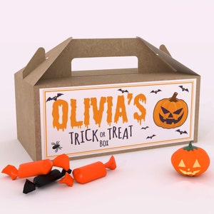 Puede incluir: Una caja de cartón marrón con asa, decorada con un tema de Halloween. La caja tiene una etiqueta que dice "Olivia's Trick or Treat Box" con una calabaza y murciélagos. También hay algunos caramelos envueltos en naranja y negro delante de la caja.