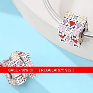 May include: A silver charm bead in the shape of a cube, adorned with hearts, squares, and sparkling accents. The cube features red, pink, and cream-colored squares. The charm is designed to be strung on a bracelet or necklace.