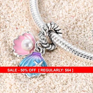 May include: A silver charm bracelet with two seashell charms. One charm is a pink and white seashell with a pearl, and the other is a blue and purple seashell with a star. The bracelet is on a sandy surface.