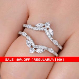 May include: A sparkling, silver-toned ring with a double band design. The bands are adorned with numerous clear, round-cut and marquise-cut gemstones, creating a delicate, leaf-like pattern. The ring is set against a skin-toned background.