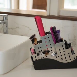 May include: A silver metal organizer with a black base, designed to hold various bathroom items. It features a pink hair straightener, a purple hair brush, and other toiletries.