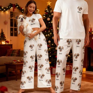 May include: Matching white pajama sets with a dog face print and the name "Bella". The sets include a short-sleeved crop top and wide-leg pants for the woman, and a t-shirt and pants for the man. The dog in the print is a small, fluffy breed.