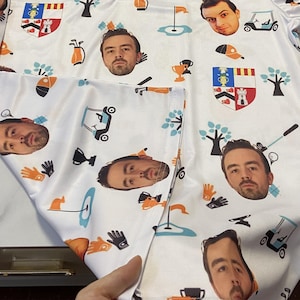 May include: White golf shirt featuring a repeating pattern of a man's face, golf-related graphics, and heraldic crests. The design includes golf carts, gloves, trees, and golf flags. The shirt is made of a lightweight material.