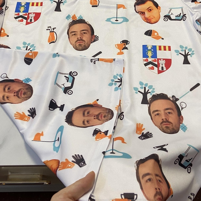 May include: White golf shirt featuring a repeating pattern of a man's face, golf-related graphics, and heraldic crests. The design includes golf carts, gloves, trees, and golf flags. The shirt is made of a lightweight material.