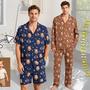 May include: Two pajama sets are displayed. One is navy blue with a pattern of baby faces, stars, and the text "Best Dad Ever." The other is brown with the same pattern. The sets include a button-down shirt and shorts or pants.
