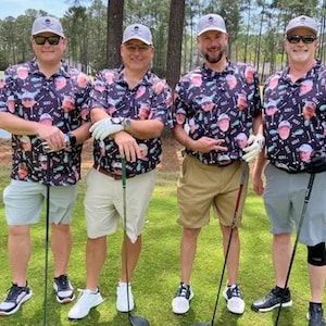 May include: Four men wearing black and white patterned golf shirts with faces printed on them. The shirts have the word "golf" printed on them. The men are holding golf clubs and are standing on a green grassy area.