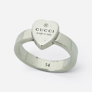 May include: A silver-toned metal ring featuring a heart-shaped centerpiece. The heart is engraved with the text GUCCI, a registered trademark symbol, and the words made in italy. The number 14 is clearly engraved on the inner band of the ring.