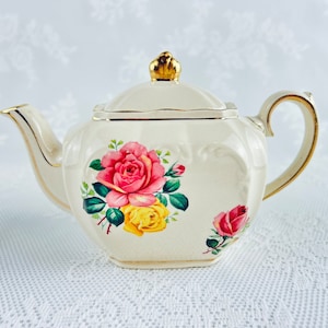 May include: A white teapot with a gold lid and handle. The teapot is decorated with pink, yellow, and red roses.