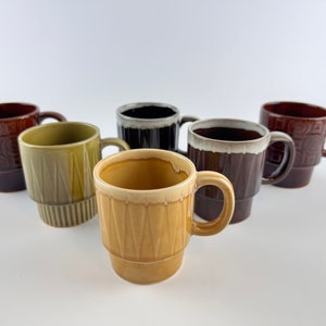 May include: Six ceramic coffee mugs in various colours and patterns. The mugs are stacked on top of each other. The mugs are all different sizes and shapes.