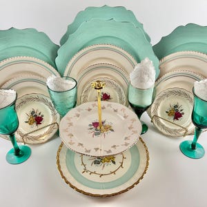 May include: A two-tiered cake stand with a gold metal base and a teal and white floral design. The stand is surrounded by teal chargers, white dinner plates, and teal glassware.