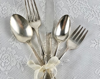 May include: A set of silver cutlery, including a knife, fork, and two spoons, tied together with a white ribbon. The cutlery is arranged on a white lace tablecloth.
