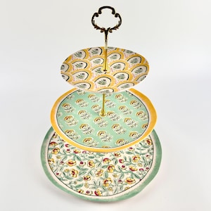 May include: A three-tiered serving tray with a decorative handle. Each tier has a different floral pattern in yellow, green, and cream. The tray is suitable for displaying desserts or appetizers. The handle is a dark bronze colour.