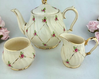 May include: A white teapot, creamer, and sugar bowl set with a gold trim and pink rose pattern. The teapot has a lid with a gold knob.