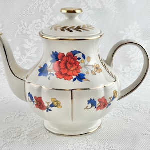 Peut inclure: Théière blanche avec une bordure dorée et un motif floral. Les fleurs sont rouges, bleues et jaunes. La théière a un couvercle avec un bouton doré.