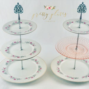 May include: Two three-tiered cake stands with decorative floral plates and a pink dotted plate. The stands are silver and have a decorative finial on top. The text "pretty plates curated" is visible in the background.