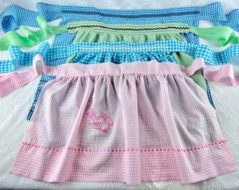 Op de afbeelding: Drie gingham schorten met blauwe, groene en roze geruite patronen. De roze schort heeft een klein geborduurd vogeltje.