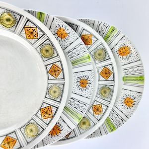 May include: A set of white ceramic plates with a geometric design. Each plate has a repeating pattern of orange, yellow, and black squares and circles. The plates have a decorative border with green and black stripes.