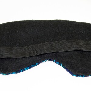 K&ouml;nnte beinhalten: Schwarze Schlafmaske mit elastischem Band. Die Maske ist mit schwarzem Stoff gef&uuml;ttert und hat eine blaue und t&uuml;rkisfarbene Vorderseite.