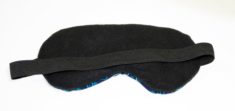 K&ouml;nnte beinhalten: Schwarze Schlafmaske mit elastischem Band. Die Maske ist mit schwarzem Stoff gef&uuml;ttert und hat eine blaue und t&uuml;rkisfarbene Vorderseite.
