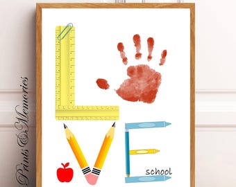 May include: A framed print with the word "LOVE" spelled out using school supplies. The "L" is a yellow ruler with a paperclip on top, the "O" is a red handprint, the "V" is two yellow pencils, and the "E" is three blue crayons. The word "school" is written below the crayons.