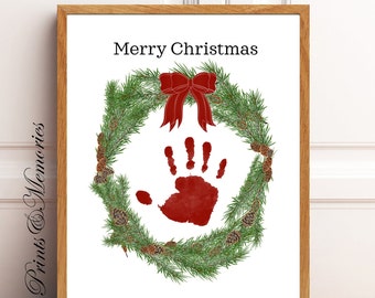 Puede incluir: Una impresión enmarcada con temática navideña. La obra presenta una corona verde con piñas, una huella de mano roja y un lazo rojo. Las palabras "Merry Christmas" están impresas en la parte superior. El marco es de madera marrón claro.